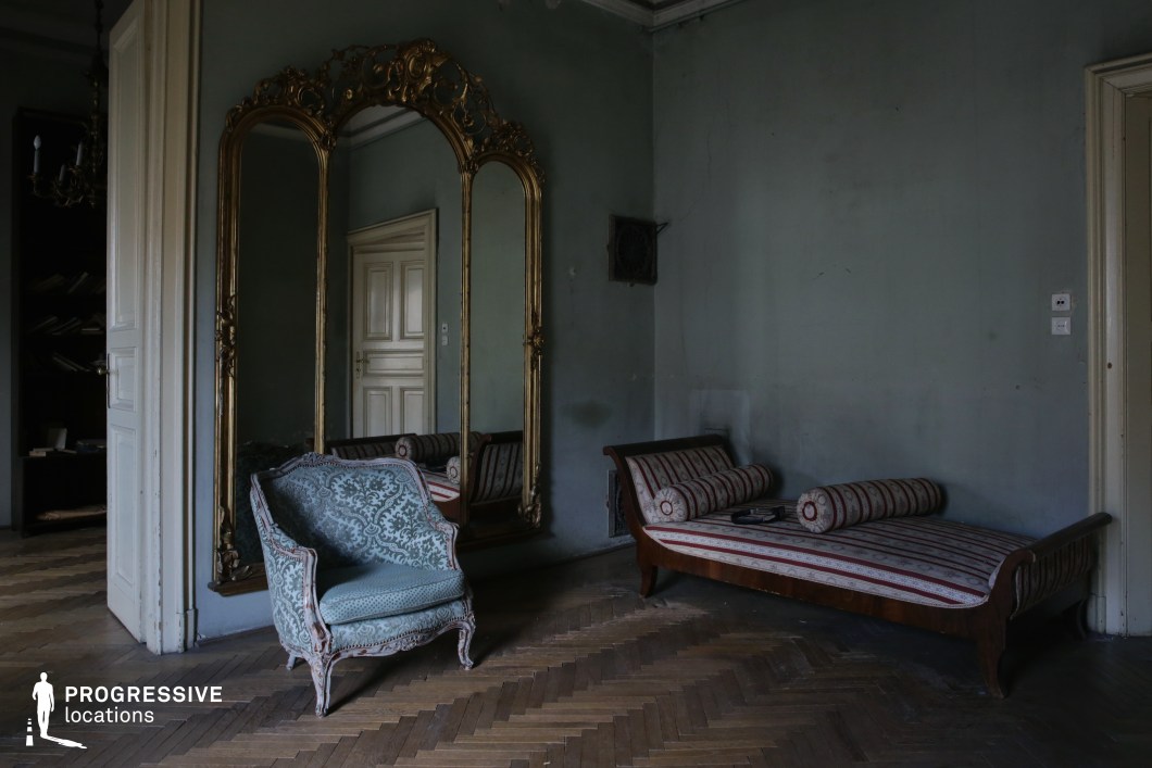 This aged interior exudes a sense of faded grandeur, featuring an opulent gold-framed mirror, distressed pastel-green walls, and antique furniture, including a striped chaise lounge and an intricately patterned armchair. The wooden parquet flooring, high ceilings, and paneled doors suggest a once-luxurious residence that has been left to time. The dim lighting and worn textures enhance its melancholic yet elegant atmosphere. This location is ideal for period dramas, psychological thrillers, or introspective character-driven narratives, offering a visually rich setting for scenes depicting nostalgia, solitude, or the remnants of aristocratic wealth.