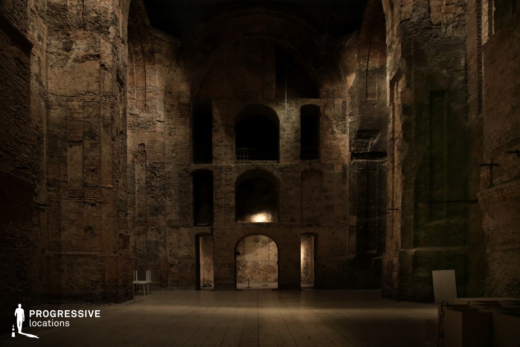 This image presents an expansive, dimly lit interior space with towering red brick walls, arched openings, and aged architectural details. The worn surfaces and deep recesses in the structure create an atmosphere of mystery and historical weight. The grand scale of the room is emphasized by its soaring ceilings and dramatic lighting, which casts shadows across the textured walls. The sparse furnishings add to the sense of abandonment and raw authenticity. This location is ideal for historical epics, fantasy films, or gothic thrillers, serving as a backdrop for secret meetings, otherworldly rituals, or medieval-inspired storytelling.