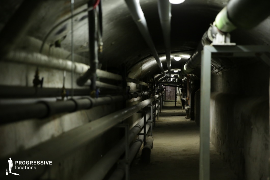 The image captures a narrow, dimly lit underground tunnel lined with an intricate network of exposed pipes running along both walls and the ceiling. The curved, concrete enclosure creates a confined, claustrophobic atmosphere, while overhead fluorescent lighting casts a cold, utilitarian glow. The uneven dirt floor and signs of wear on the infrastructure suggest an aging or neglected industrial space. The linear perspective enhances the sense of depth and secrecy, making this location ideal for suspenseful thrillers, dystopian sci-fi, or espionage films, particularly for chase sequences, covert escapes, or tension-filled explorations of hidden facilities.