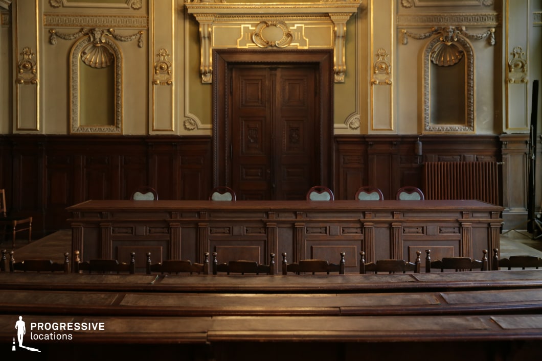 This stately courtroom exudes authority and historical elegance, featuring dark wood paneling, carved furnishings, and gilded architectural detailing. The high-backed judges' bench sits against an imposing wooden door framed by intricate moldings and decorative arches. The symmetrical layout, with polished wooden desks and tiered seating, reinforces the room’s solemn and institutional character. The warm, muted lighting and refined craftsmanship contribute to a dignified yet imposing atmosphere. This location is ideal for legal dramas, political thrillers, or historical narratives, offering a distinguished setting for courtroom battles, high-profile trials, or secretive power struggles.