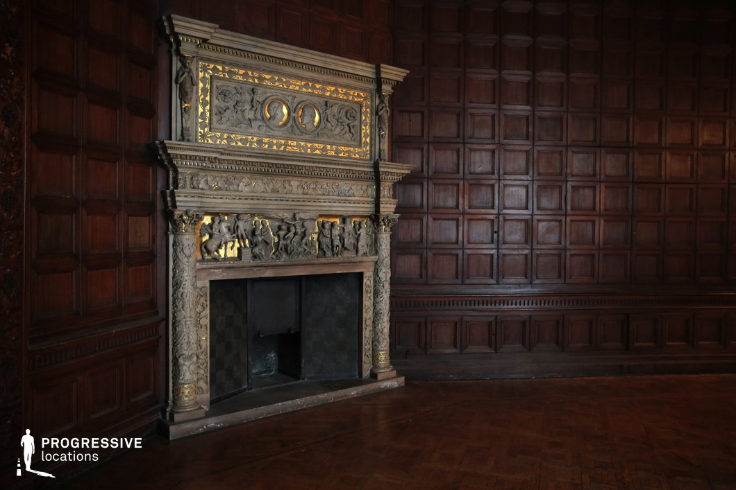 This stately interior features dark wood-paneled walls and an elaborately carved fireplace adorned with intricate sculptural reliefs and gilded detailing. The craftsmanship of the fireplace, with its mythological figures and classical motifs, stands as the room’s focal point, exuding historical opulence. The dimly lit ambiance and rich textures enhance the sense of grandeur and secrecy. This location is ideal for period dramas, historical thrillers, or gothic mysteries, providing an atmospheric setting for aristocratic gatherings, clandestine meetings, or moments of introspection in a setting that evokes power and old-world prestige.