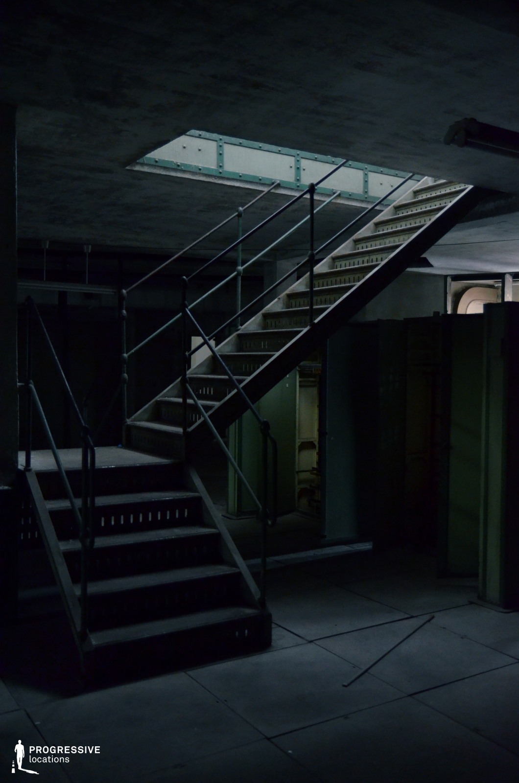 This industrial-style metal staircase, featuring dark railings and perforated steps, ascends through a raw, concrete interior. The exposed structural elements, including steel beams and bolted reinforcements, emphasize the utilitarian nature of the space. The surrounding walls are lined with aged metal panels and cabinets, hinting at a mechanical or power facility setting. The dim lighting, with a single opening casting a faint glow from above, creates an atmospheric interplay of shadows and highlights. This location is ideal for tense, suspenseful scenes in thriller, sci-fi, or post-apocalyptic films, offering a confined, industrial aesthetic that enhances a sense of mystery or isolation.