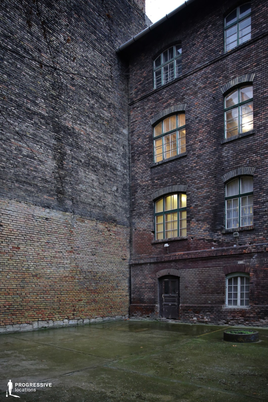 The image depicts a secluded courtyard enclosed by tall, aged red brick walls with arched windows framed in green metal. The uneven brickwork, patches of discoloration, and worn surfaces contribute to a weathered, industrial aesthetic. The ground is covered in damp concrete, adding to the raw, unpolished atmosphere. A small, dark wooden door at the base of the building suggests a hidden or restricted space. The setting evokes a sense of isolation and secrecy, making it ideal for crime dramas, urban thrillers, or neo-noir films, particularly scenes involving covert meetings, escapes, or clandestine activities.