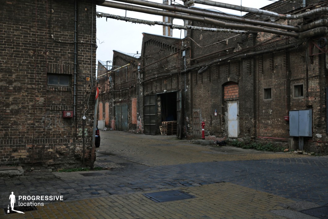 The image depicts a desolate industrial alley lined with weathered red brick factory buildings, featuring arched windows, rusted metal doors, and patches of exposed brickwork. Overhead, a tangle of corroded pipes runs across the alley, adding to the gritty, decayed atmosphere. The cobblestone and brick-paved ground is uneven, showing signs of wear, while an open warehouse door hints at lingering industrial activity. The location has a raw, cinematic quality, making it ideal for crime thrillers, post-apocalyptic settings, or noir-style films, serving as a perfect backdrop for clandestine meetings, illicit operations, or high-stakes chases through abandoned industrial zones.