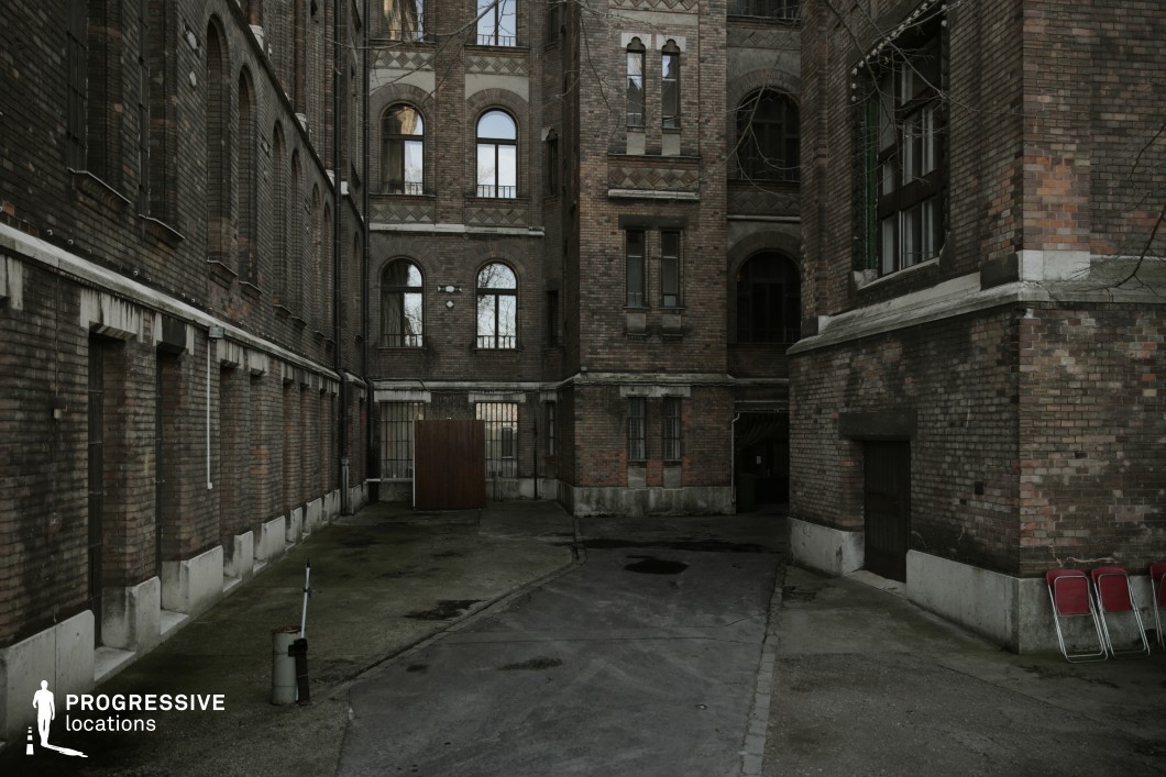 This image depicts a confined inner courtyard surrounded by towering red brick buildings with arched windows and intricate decorative patterns. The aged facades, with their darkened surfaces and barred windows, evoke a sense of history and secrecy. The narrow, uneven pavement, small puddles, and scattered objects contribute to the overall worn and slightly ominous atmosphere. The enclosed layout creates a sense of isolation, making this location ideal for noir crime films, psychological thrillers, or historical dramas. The dramatic verticality and shadowed entryways provide a cinematic setting for tense confrontations, secretive exchanges, or moments of suspenseful observation.