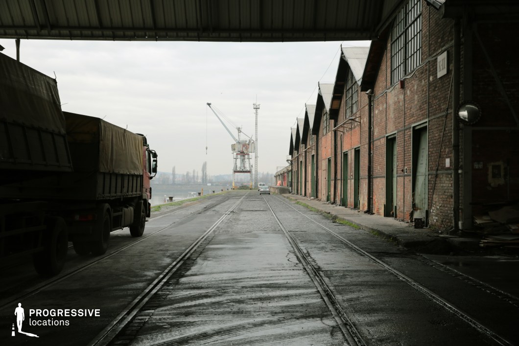 The image depicts a long stretch of red-brick warehouses with green metal doors, aligned along a riverside loading dock. Embedded railway tracks run parallel to the structures, emphasizing the location's historical function as a transportation hub. A covered loading area frames the foreground, partially concealing a parked cargo truck, while in the distance, a large crane looms over the water, reinforcing the site's industrial character. The damp pavement and worn infrastructure contribute to a gritty, utilitarian aesthetic. This location is ideal for crime dramas, neo-noir thrillers, or action films, providing an evocative setting for smuggling operations, clandestine deals, or high-stakes chases along the waterfront.