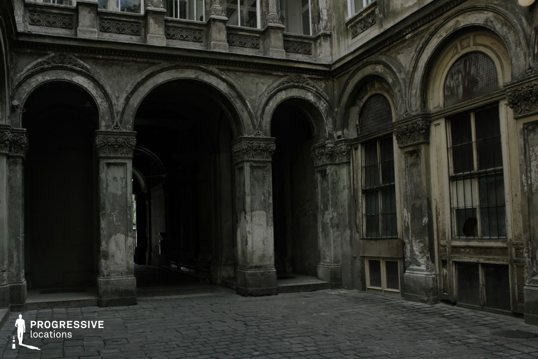 This image captures an abandoned inner courtyard surrounded by elaborate yet deteriorating architecture. The grand arched passageways, adorned with intricate carvings, lead into shadowy corridors, emphasizing depth and mystery. The peeling paint, cracked plaster, and barred windows add to the aged, forgotten atmosphere, suggesting a past grandeur that has since faded. The cobblestone ground enhances the historical feel, while the dimly lit interior archways create a strong visual contrast with the open courtyard. This location is ideal for historical dramas, gothic horror, or noir thrillers, serving as a hauntingly beautiful backdrop for secretive encounters, chase sequences, or explorations of forgotten places.
