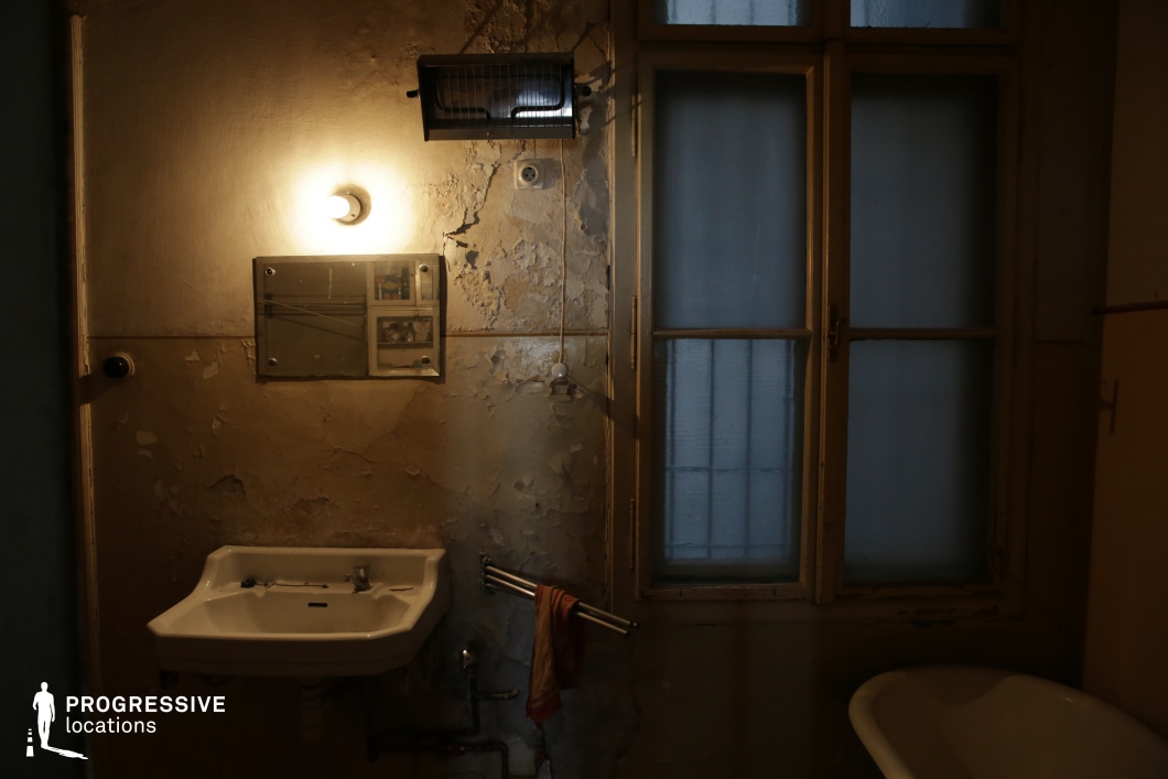 This aged bathroom exudes a sense of neglect and decay, with peeling paint, exposed plumbing, and dim, uneven lighting casting shadows across the worn surfaces. The frosted glass window, framed by deteriorating wooden panels, allows only a faint glow to seep in, reinforcing the confined and eerie atmosphere. The old ceramic sink, rusting fixtures, and a solitary hanging cloth add to the sense of abandonment. This location is ideal for psychological thrillers, horror films, or neo-noir crime dramas, offering a raw and unsettling visual space that enhances tension, secrecy, and psychological unease.
