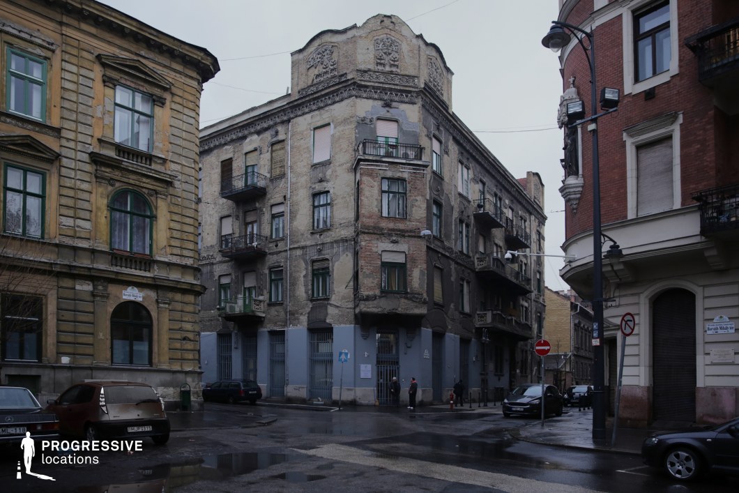 This image captures a worn-out corner building with peeling plaster, exposed brickwork, and small wrought-iron balconies, standing at the intersection of a historic yet decayed urban district. The surrounding architecture, featuring intricate facades with faded grandeur, suggests a once-elegant neighborhood that has fallen into neglect. The damp streets, scattered puddles, and muted color palette enhance the sense of urban decay. The heavy security gates on the ground level add an element of restriction, reinforcing the area’s gritty, lived-in character. This location is ideal for crime dramas, neo-noir thrillers, or social realism films, providing a compelling backdrop for stories of struggle, hidden histories, or illicit dealings in the margins of the city.