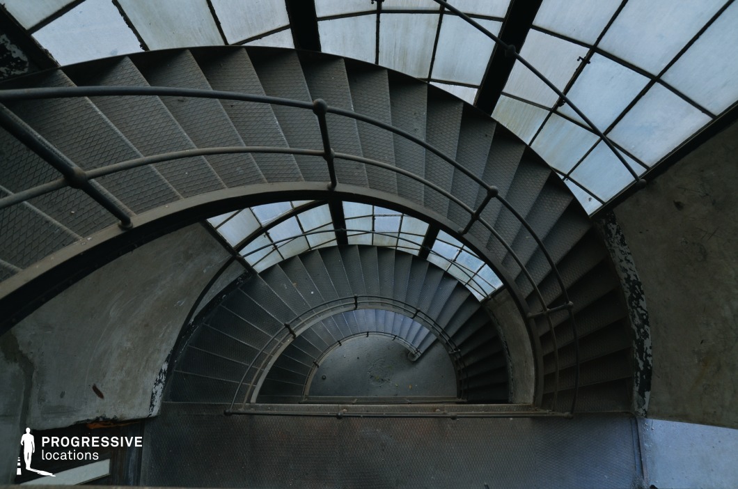 The image captures a dramatic downward view of a spiral staircase with metal steps and railings, enclosed by a curved glass canopy. The industrial-style design features textured steel treads and a utilitarian handrail, creating a sense of depth and movement. The arched glass panels above allow diffused light to filter through, casting a cool-toned ambiance over the worn concrete walls. The geometric repetition of the steps and railings enhances the visual dynamism, making this location ideal for psychological thrillers, noir films, or action sequences involving pursuits, escapes, or moments of suspense.