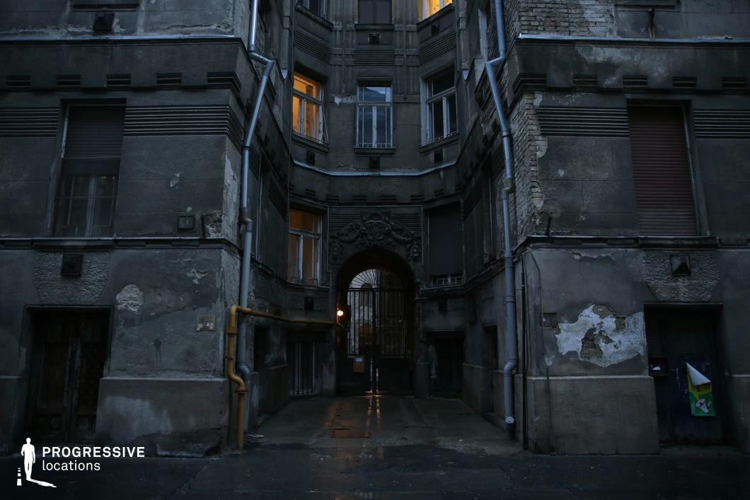 This image captures a dark, enclosed courtyard surrounded by weathered buildings with crumbling facades, peeling plaster, and exposed brickwork. The central feature is an ornate archway with a heavy metal gate, partially illuminated by a dim interior light beyond. The damp pavement reflects the subtle glow from scattered windows, adding depth to the cold, desolate atmosphere. Rusted pipes, boarded-up windows, and architectural details hint at a forgotten grandeur now overtaken by neglect. This location is ideal for neo-noir thrillers, psychological dramas, or crime films, serving as an ominous backdrop for secretive dealings, hidden pasts, or scenes of urban tension and isolation.