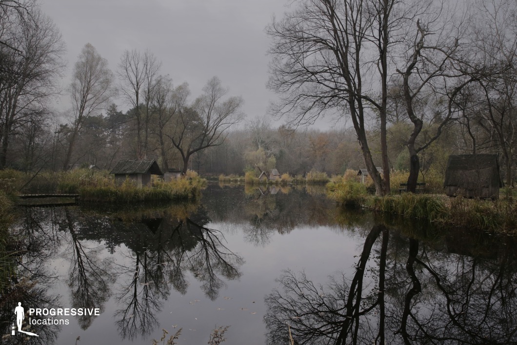 The image showcases a tranquil yet eerie lakeside landscape, where small, rustic huts are scattered along the shore, surrounded by tall, bare trees and patches of reeds. The calm water creates an almost perfect mirror effect, reflecting the trees and sky, enhancing the surreal and dreamlike quality of the scene. The distant tree line and misty atmosphere contribute to a sense of solitude and remoteness. This location is ideal for psychological thrillers, horror films, or contemplative dramas, offering a hauntingly beautiful backdrop for themes of isolation, mystery, or forgotten places.