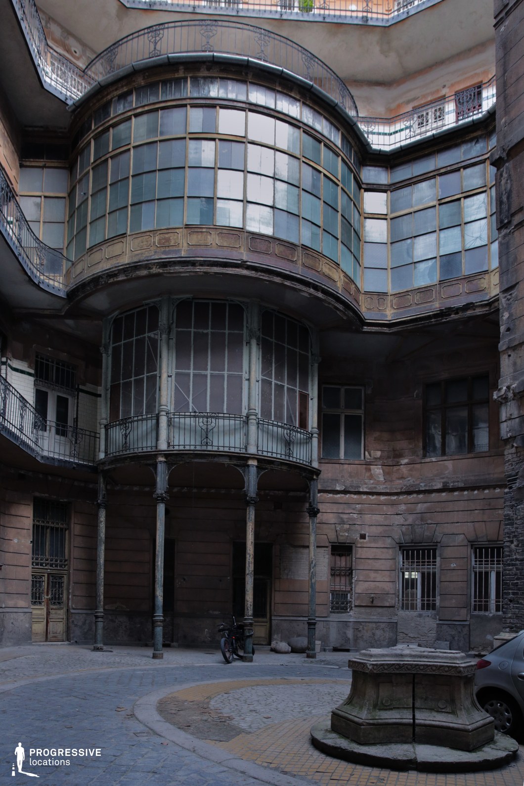 This image presents a decayed yet once-elegant inner courtyard of a historic tenement building, featuring an imposing curved glass bay window supported by delicate iron columns. The worn facade, with peeling plaster and rusted railings, hints at a past grandeur that has faded over time. The cobblestone ground, barred windows, and the presence of an old well-like structure at the center add to the location’s nostalgic and melancholic atmosphere. This setting is ideal for noir thrillers, period dramas, or mystery films, providing a visually rich backdrop for scenes of intrigue, forgotten histories, or clandestine meetings in an overlooked part of the city.