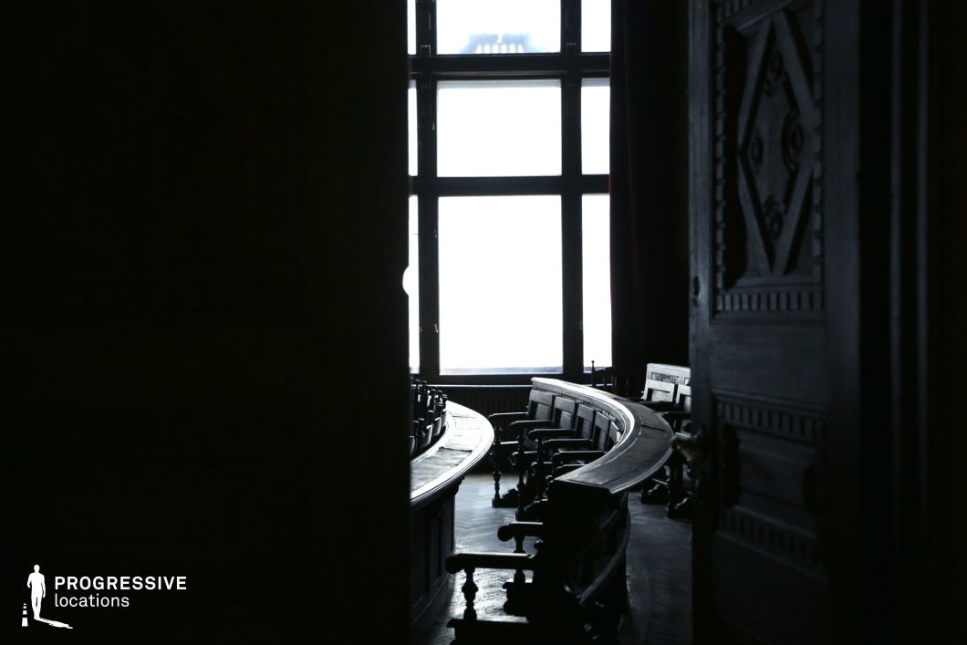 This shadowed, wood-paneled courtroom exudes a sense of authority and secrecy, with its curved rows of aged wooden benches partially illuminated by a large, backlit window. The slightly ajar ornate door in the foreground adds an element of intrigue, suggesting entry into a space of judgment and consequence. The contrast between darkness and the overexposed window enhances the moody, dramatic atmosphere. This location is ideal for legal dramas, political thrillers, or noir-style storytelling, serving as a striking backdrop for intense courtroom debates, covert negotiations, or morally complex deliberations.