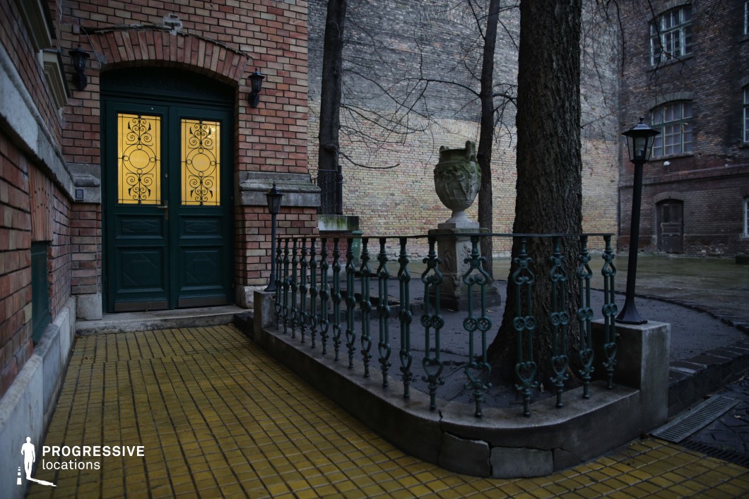 This enclosed courtyard blends elegance with a sense of quiet abandonment, featuring weathered brick facades, an iron-railed platform, and a grand green double door with intricate wrought iron detailing. The warm glow from the glass panels contrasts with the muted tones of the aged brick and stone surfaces. Decorative elements, such as a sculpted stone urn on a pedestal and vintage-style lampposts, enhance the location's old-world charm. The cracked pavement and overgrown details contribute to a melancholic yet atmospheric setting. This location is ideal for period dramas, mystery thrillers, or gothic-inspired storytelling, providing a visually rich backdrop for scenes of secrecy, historical intrigue, or quiet reflection.