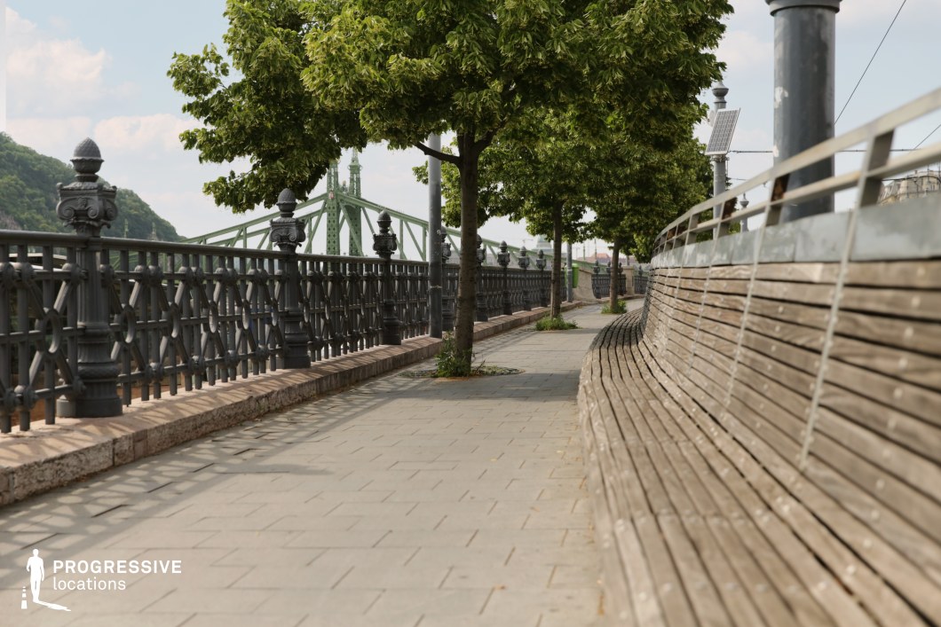 Tree-lined riverside walkway with long bench and metal railing used for photo and film shoots