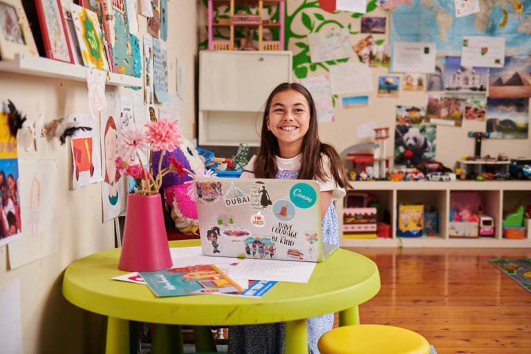 A school girl sitting in a classroom with a laptop that has stickers on it including a Canva sticker