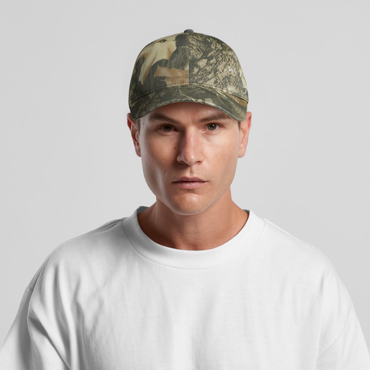 Access Camo Cap AS-1130C A camo cap featuring a mix of green and brown patterns, worn by a model in a white t-shirt. The cap has a logo.
