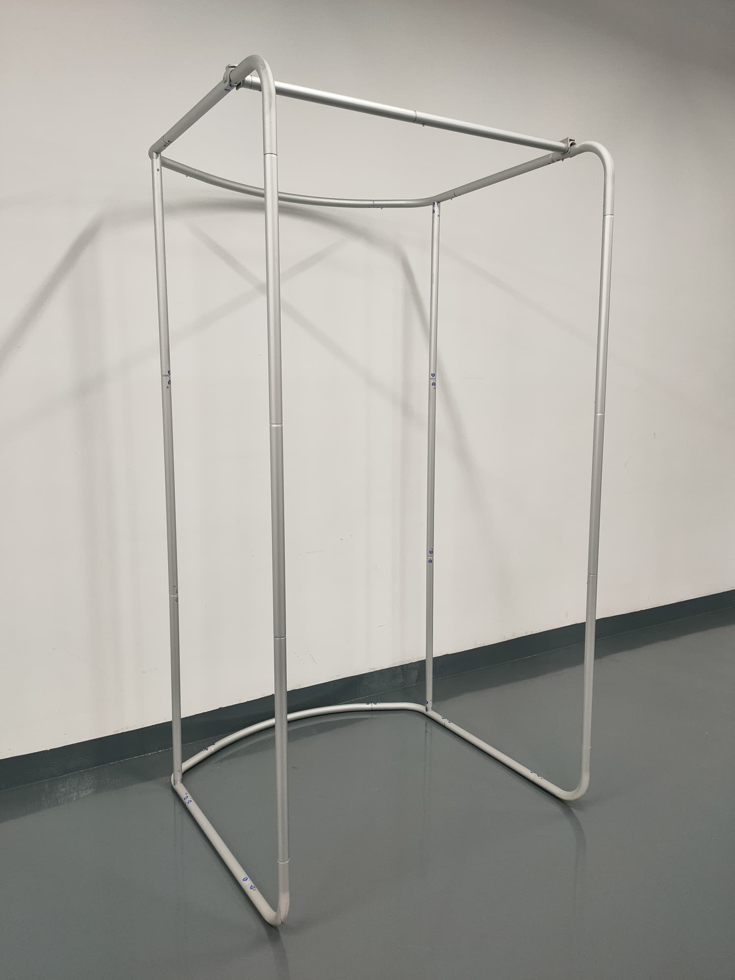 A silver tension fabric change room frame with a rectangular base and open top, set against a plain wall.