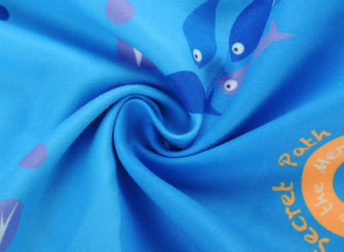 A blue beach towel with a double-faced pile featuring playful fish designs and circular motifs. It has a branding logo.