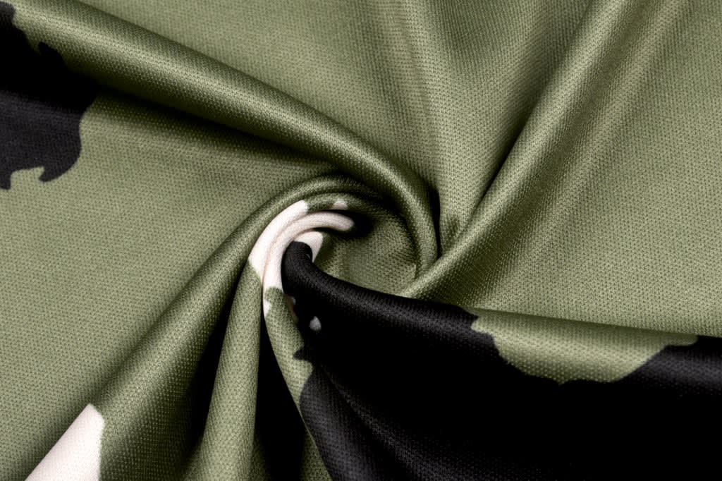 A close-up view of camouflage fabric featuring shades of green, black, and beige. The material is textured.
