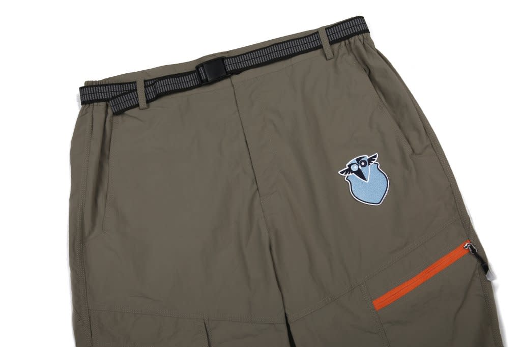 Windbreaker pants in a greenish-brown colour, featuring an orange zip pocket and a logo on the side.
