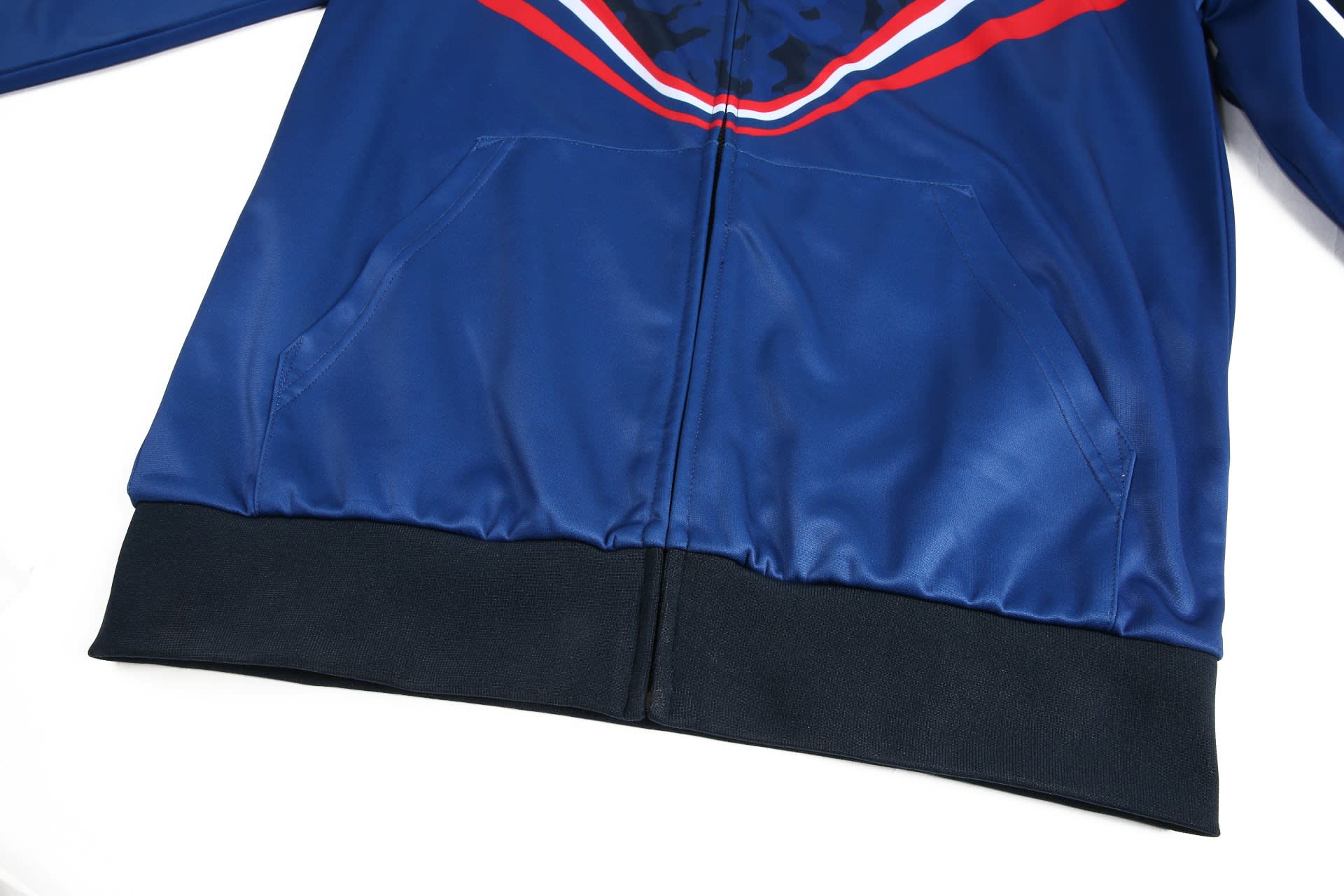A blue track suit warm-up jacket with side pockets and a contrasting black hem, featuring a logo on the front.