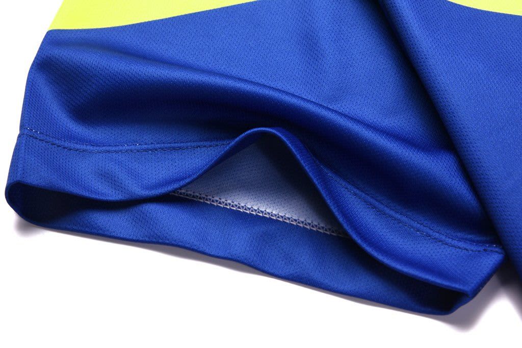 A close-up of a blue and yellow shirt, featuring a folded hem and smooth fabric texture. It has a logo present.