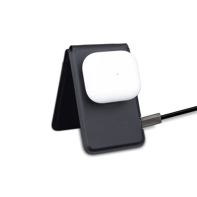 A black magnetic card wallet and wireless charging stand holding white wireless earbuds, connected by a charging cable.