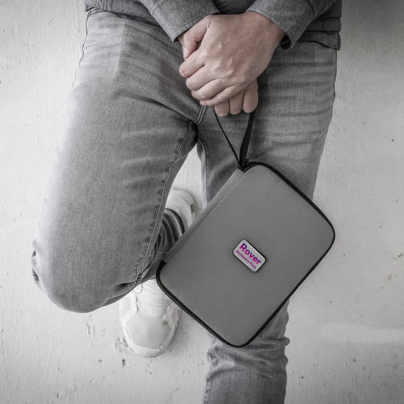A grey travel wallet with a strap, featuring a logo, held against a person's leg dressed in denim.