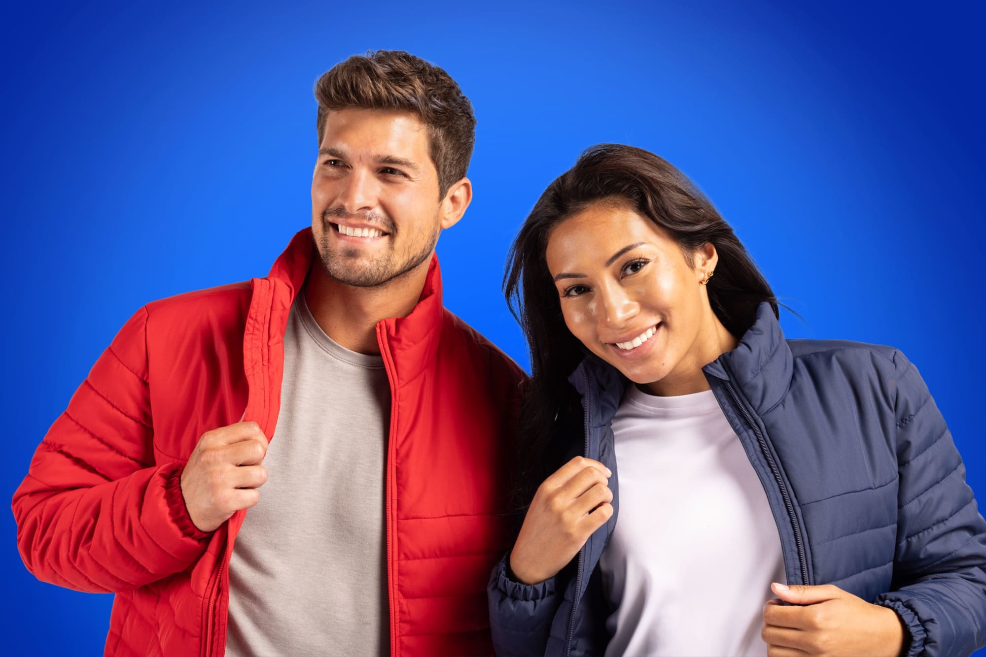 A red puffer jacket and a navy puffer jacket worn by a smiling man and woman against a blue background, both with logos.