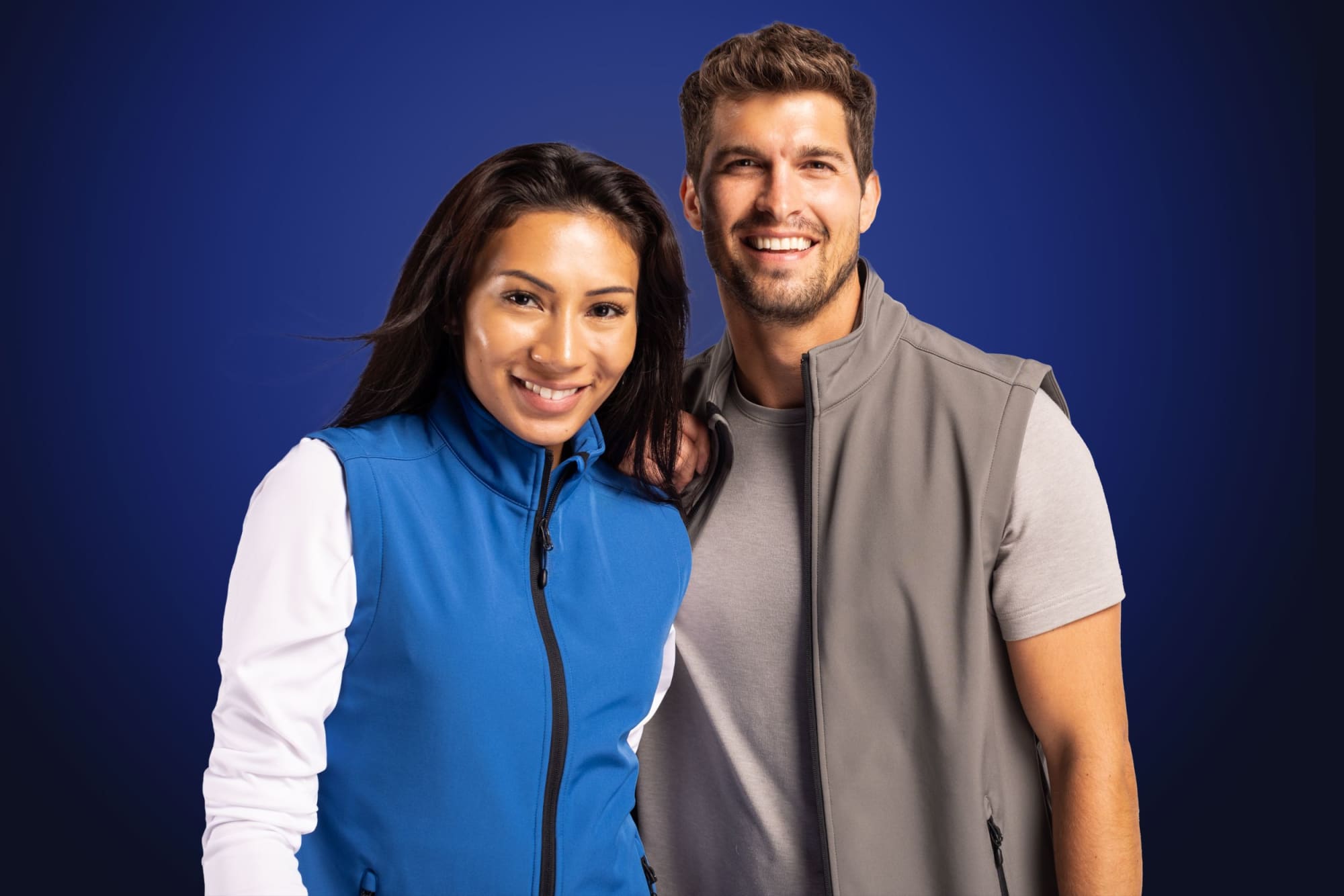 A blue eco stretch vest and a grey vest are worn by a man and a woman, both smiling. The image features branding.