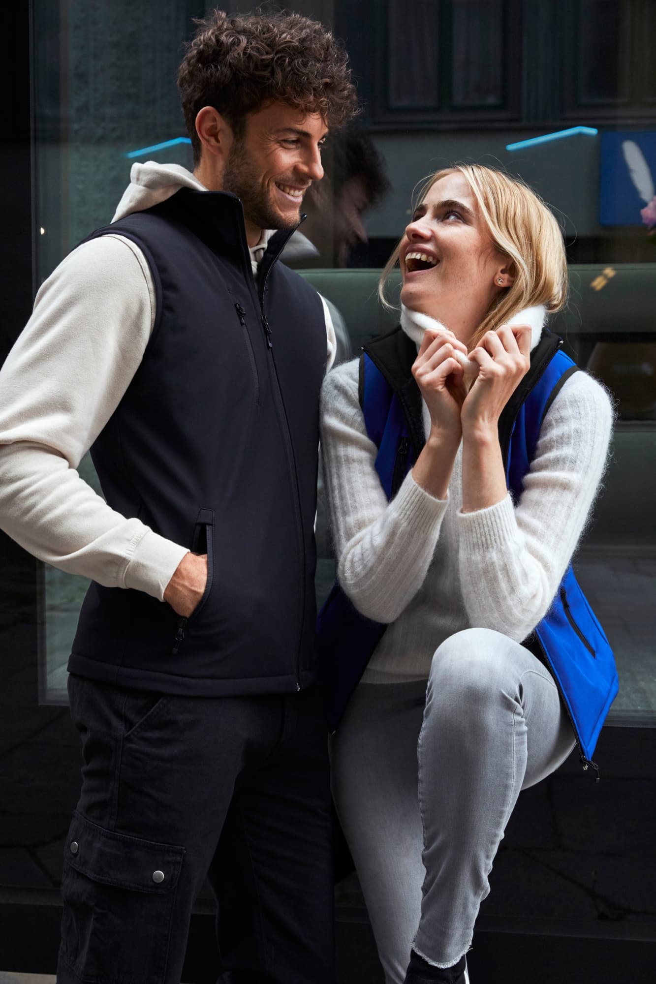 A black softshell vest worn by a man, paired with a blue softshell vest worn by a woman, both smiling.