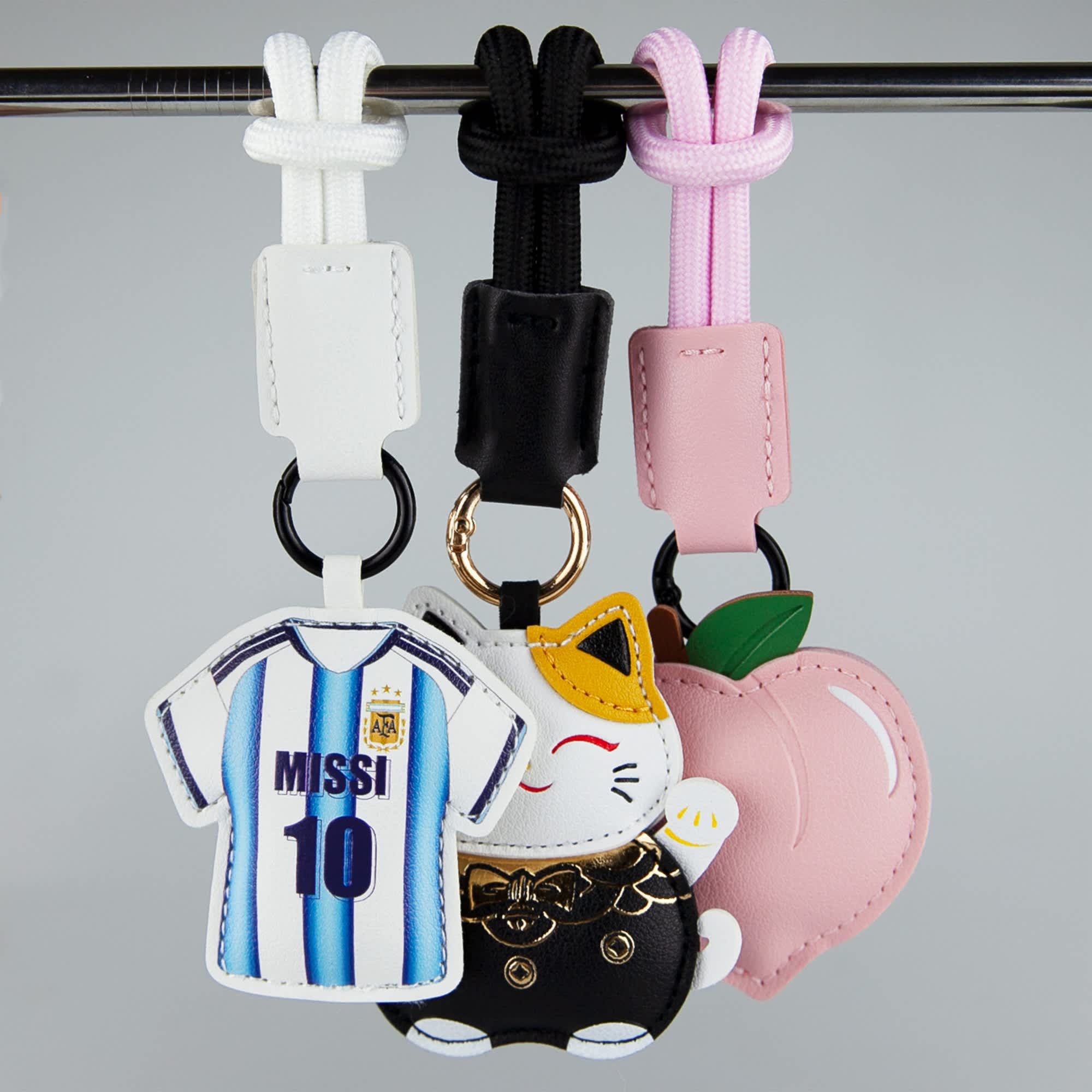 Three bag charms featuring a jersey, a cat, and a peach, in white, black, pink, and multicoloured designs.
