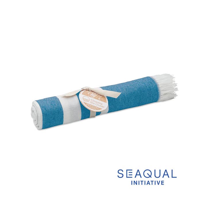 A rolled beach towel in shades of blue and white, featuring a fringed edge and a label. It has a brand logo.