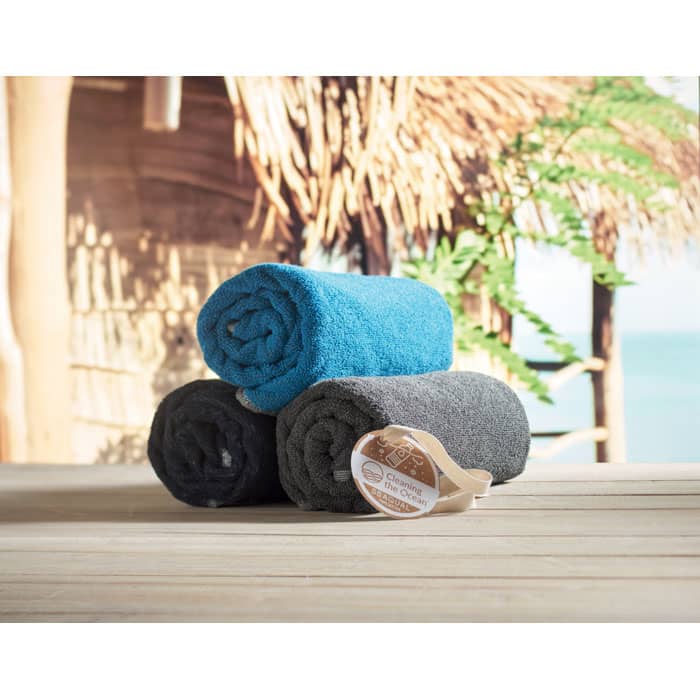A stack of three rolled towels in teal, grey, and black on a wooden surface with a tropical background.