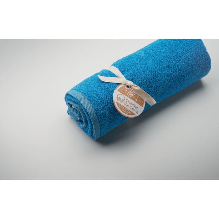 A rolled towel in a vibrant blue colour, secured with a ribbon and tag, resting on a light grey surface.