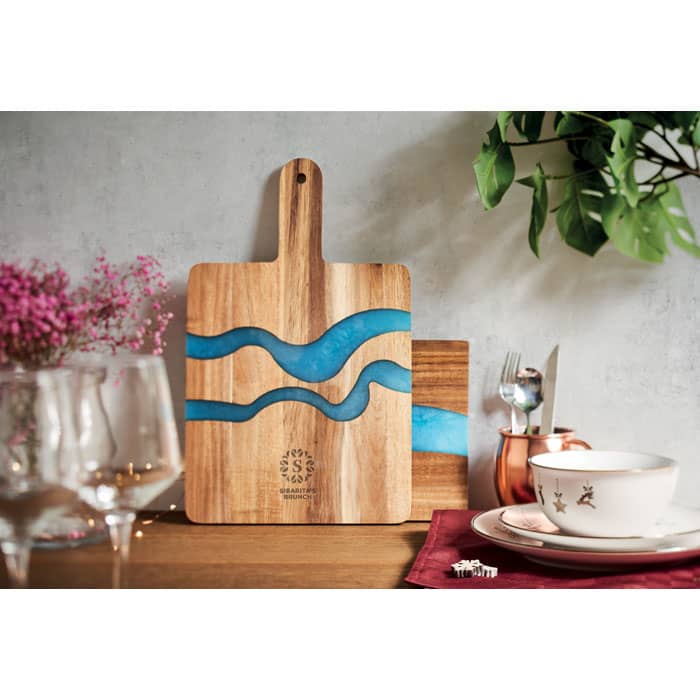 A wooden chopping board with blue resin waves, set on a table with decorative plants and dining items.