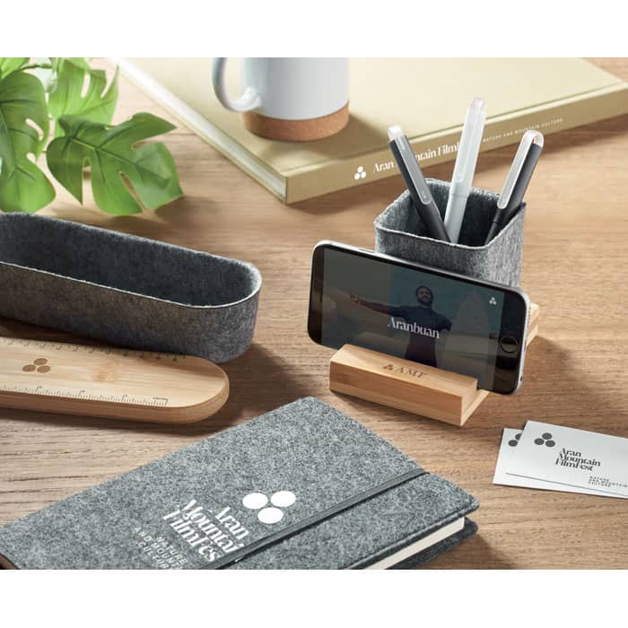 A desk setup featuring a grey felt organiser, wooden stand for a phone, notebook, and plant.