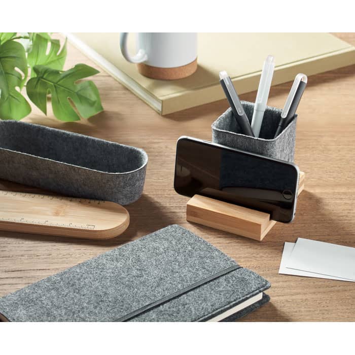 A grey felt desk organiser holds pens and a phone, alongside a notebook and bamboo ruler on a wooden desk.