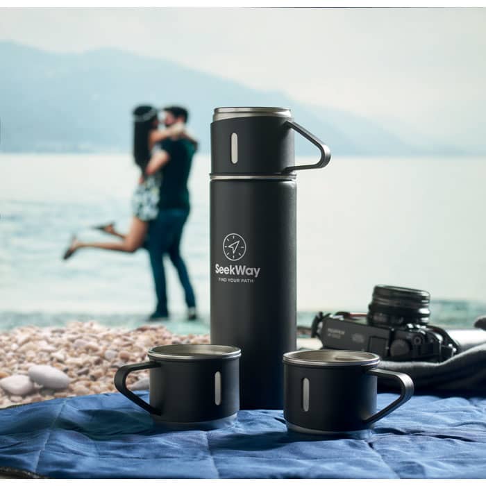 A drink bottle and two matching mugs in black, with a logo, set against a scenic outdoor background.