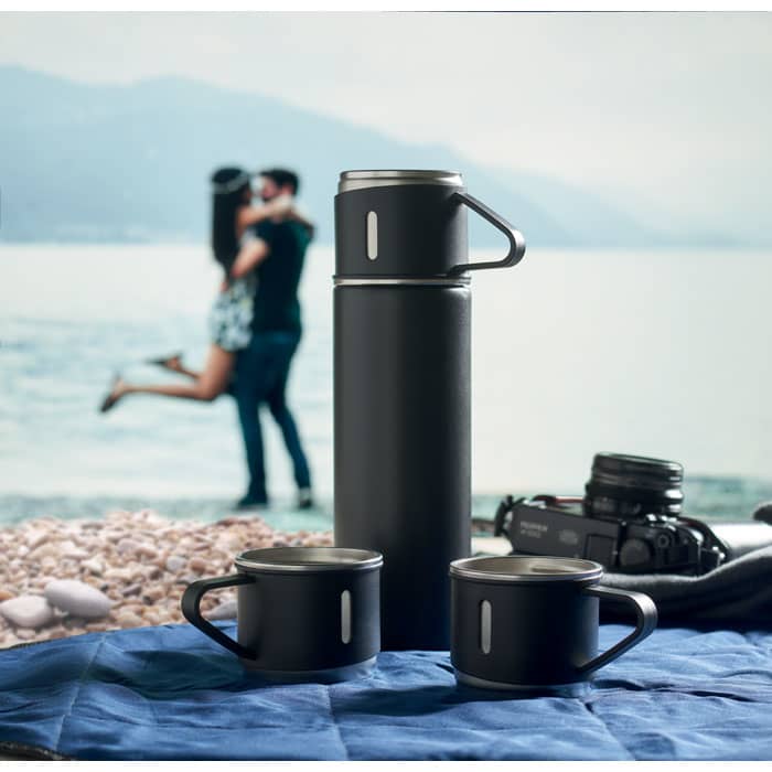 A drink bottle and two black mugs on a blue blanket by a beach, with a couple in the background.