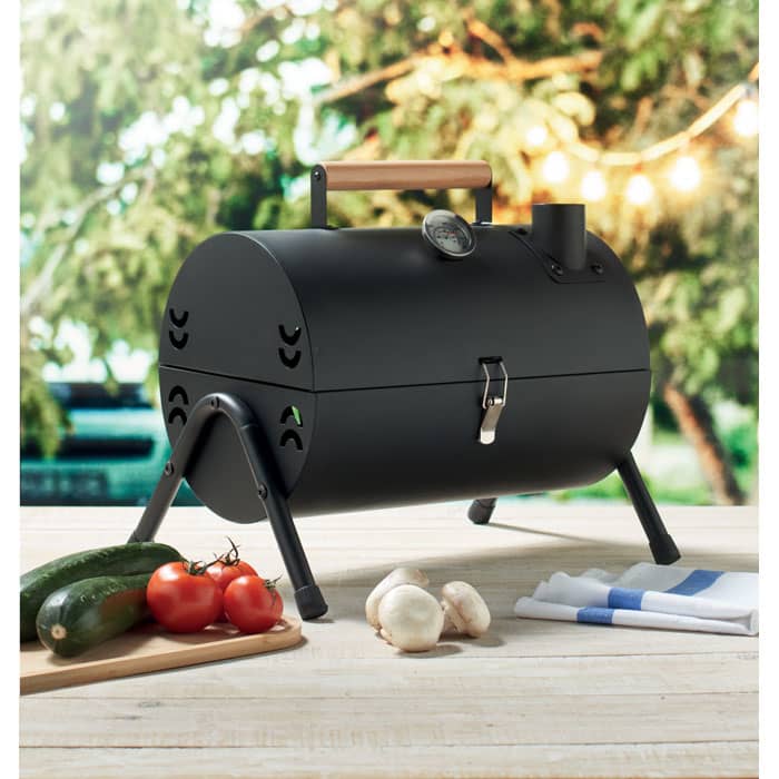 A portable black barbecue with a wooden handle, positioned beside fresh vegetables on a chopping board.