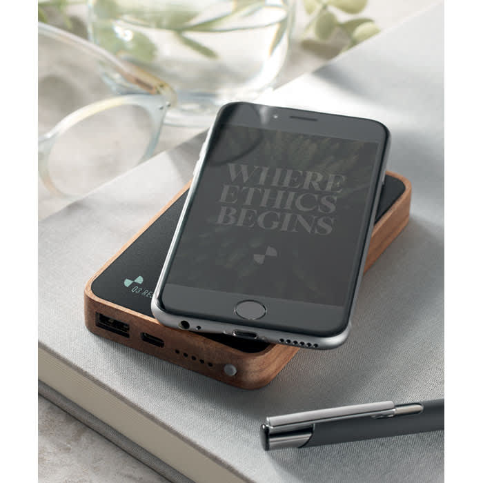 A smartphone and a sleek, wooden power bank rest on a light surface, accompanied by a pen and glasses.