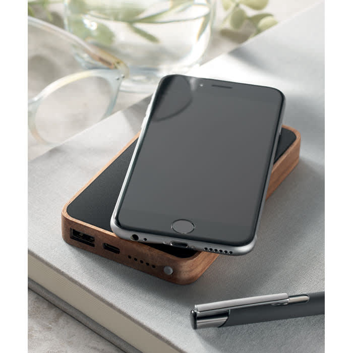 A sleek black smartphone rests on a wooden charging pad, accompanied by a pen and glasses on a grey surface.
