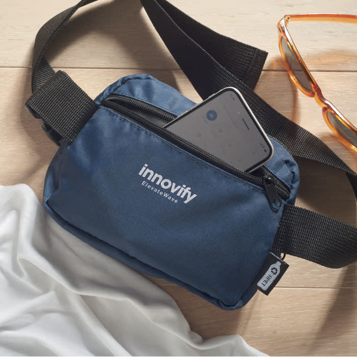 A small navy blue pouch with a zip, featuring an adjustable strap and a logo, next to glasses and fabric.