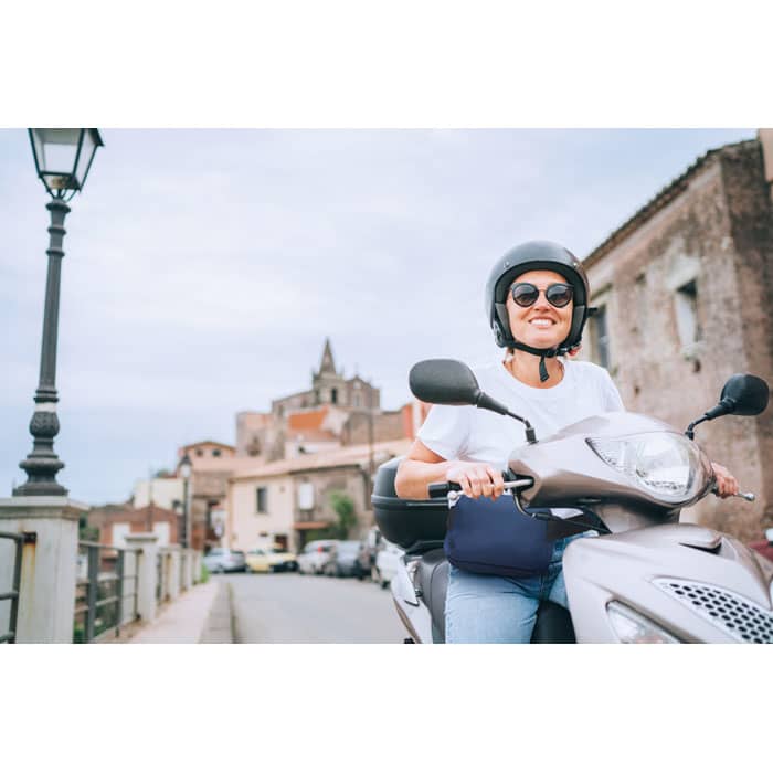 A person wearing a helmet smiles while riding a silver scooter on a street with buildings in the background.