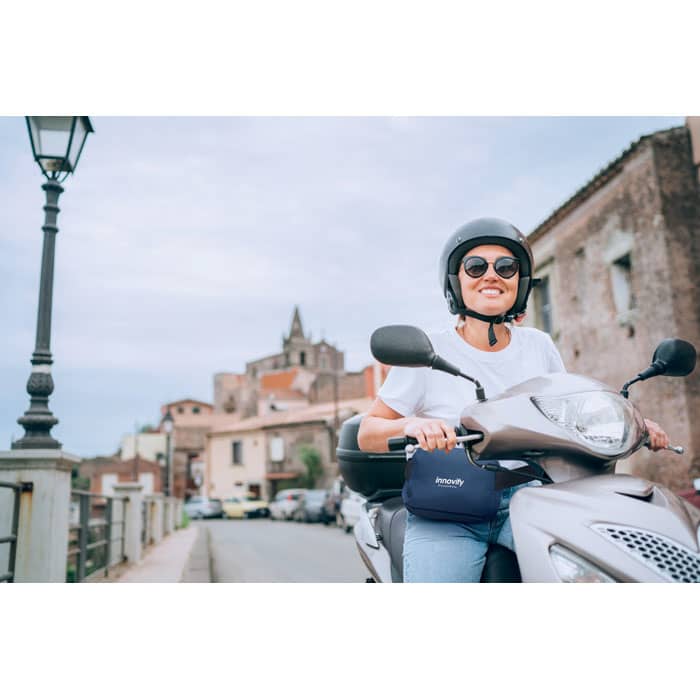 A woman wearing sunglasses and a helmet sits on a silver scooter, with a scenic town in the background.