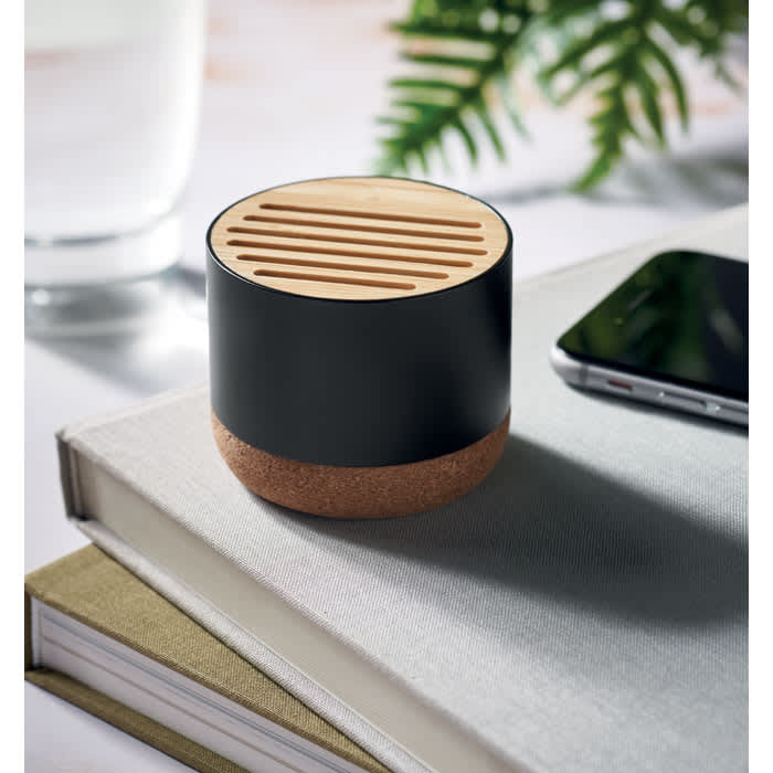 A speaker with a black top and cork base, featuring slotted design, placed on a stack of books beside a smartphone.