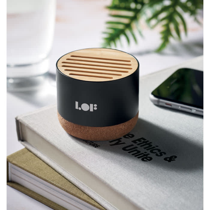 A black and cork speaker with a wooden top sits on a stack of books, alongside a smartphone and a glass of water.